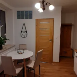 Jasne wnętrze jadalni z okrągłym stołem, trzema krzesłami, drewnianą podłogą i drzwiami. Na ścianie zegar i szklane luksfery. Widoczna szafka w tle.