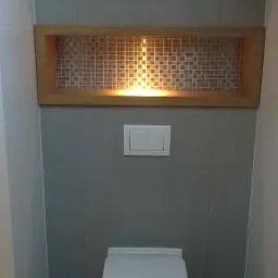 Nowoczesna toaleta podwieszana z szarymi płytkami, podświetlaną wnęką z mozaiką i skośnym sufitem.