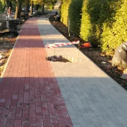Ścieżka z kostki brukowej w trakcie budowy, z wygrodzonym, niezakończonym fragmentem, czerwono-biała taśma ostrzegawcza, drzewa w tle.