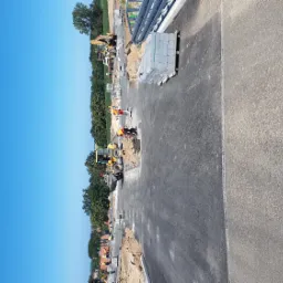 Budowa drogi z użyciem ciężkiego sprzętu i kostki. Widoczne maszyny budowlane, pracownicy w kamizelkach odblaskowych i ułożona kostka. Częściowo utwardzona nawierzchnia.