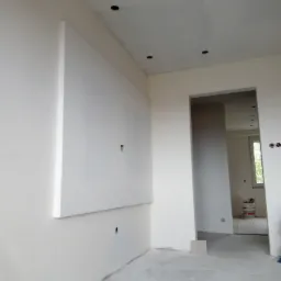 Świeżo pomalowane wnętrze z płytą karton gips na ścianie, widoczne otwory na oświetlenie w suficie, surowy beton na podłodze i gniazdko elektryczne wystające ze ściany.