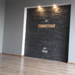 Salon po remoncie z drewnianą podłogą, szarymi ścianami i ścianą akcentującą z czarnego kamienia dekoracyjnego z wnękami na kable i oświetleniem punktowym.