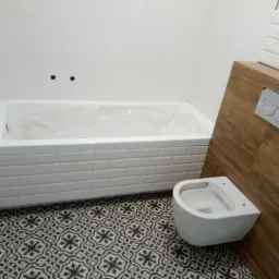 Nowoczesna łazienka z białą wanną obudowaną płytkami imitującymi cegłę, wiszącą toaletą i podłogą w czarno-białe wzory geometryczne; drewnopodobna obudowa stelaża WC z przyciskiem spłukującym.