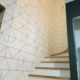 Klatka schodowa po remoncie z biało-czarną geometryczną tapetą na ścianie, drewniane stopnie schodów zabezpieczone taśmą malarską.