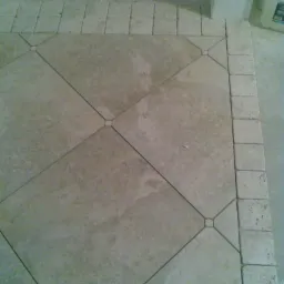 Ułożona pod kątem beżowa podłoga z płytek z ciemniejszymi fugami i obramowaniem z małych kwadratowych płytek, widoczny fragment ściany i podłogi w tle.