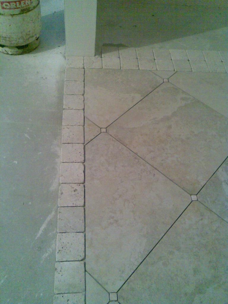 Beżowa podłoga z kwadratowych płytek ułożonych w romby z ciemniejszymi fugami, obramowana kostką brukową, widoczny fragment ściany i butla gazowa.