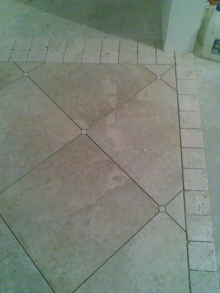 Ułożona pod kątem beżowa podłoga z płytek z ciemniejszymi fugami i obramowaniem z małych kwadratowych płytek, widoczny fragment ściany i podłogi w tle.