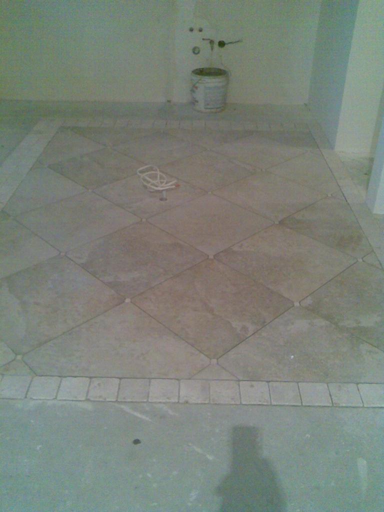 Ułożona podłoga z beżowych, kwadratowych płytek ceramicznych ułożonych w romby, z dekoracyjnym obramowaniem z mniejszych kwadratowych płytek. Widoczny fragment ściany z wyprowadzonymi instalacjami...