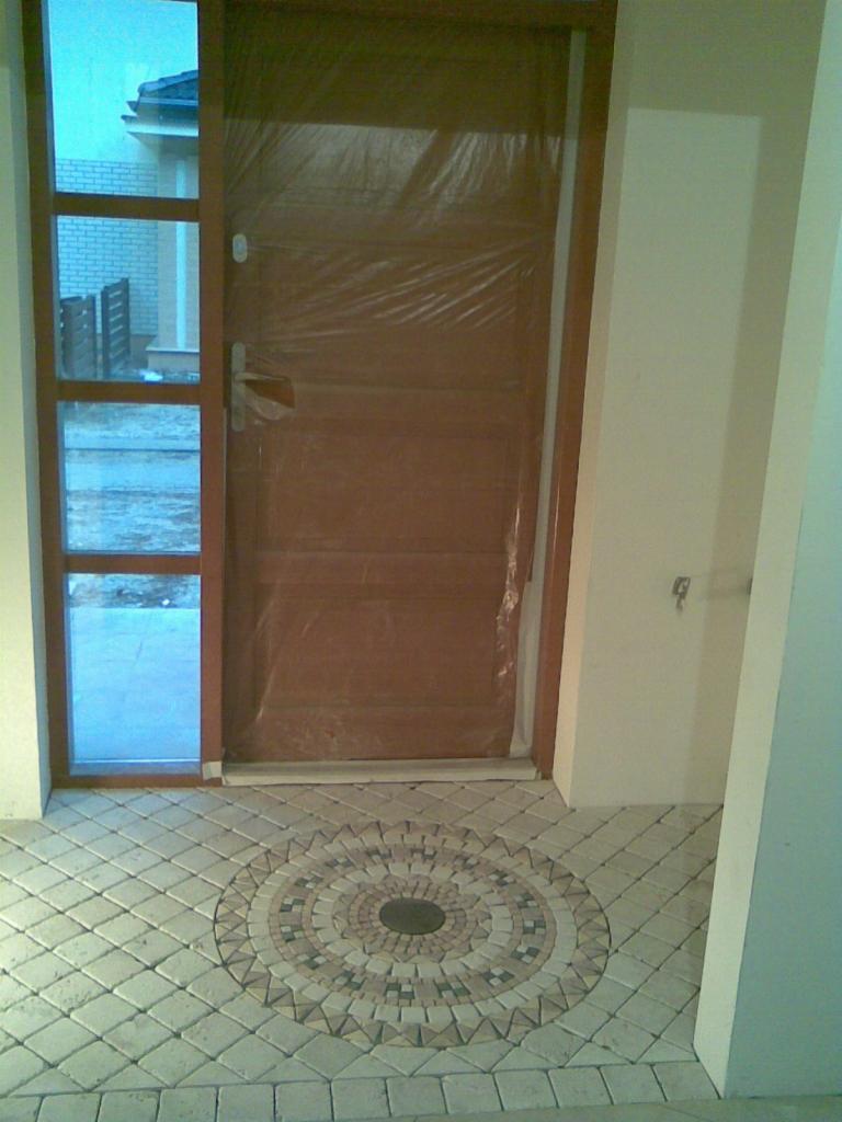 Wejście do domu z brązowymi drzwiami zabezpieczonymi folią i ozdobną mozaiką z kostki brukowej na podłodze.