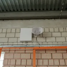 Biała tuba głośnika przymocowana do ceglanej ściany wewnątrz hali przemysłowej, obok biała skrzynka podłączeniowa, widoczne przewody i metalowa siatka ochronna z pomarańczową ramą.