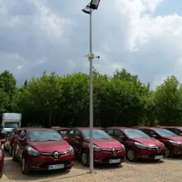 Słup oświetleniowy z kamerami monitoringu na parkingu z rzędem czerwonych samochodów osobowych i zielenią w tle, widok z dołu.