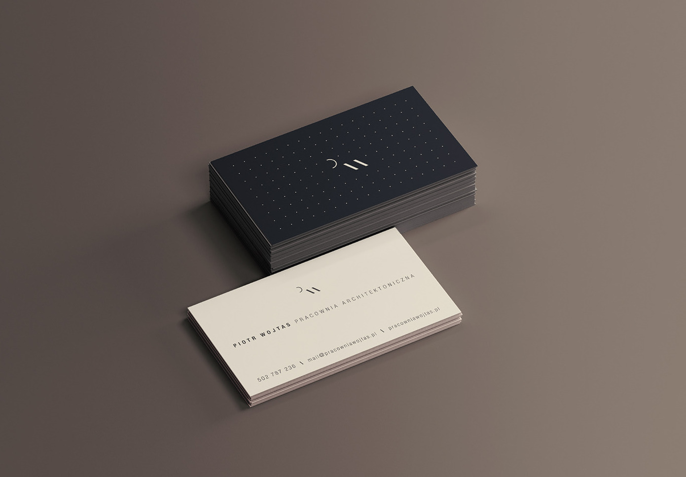 Stos wizytówek architektonicznych w odcieniach granatu i beżu, z minimalistycznym logo i danymi kontaktowymi pracowni architektonicznej Piotra Wojtasa, ułożonych na gładkim, szarym tle.