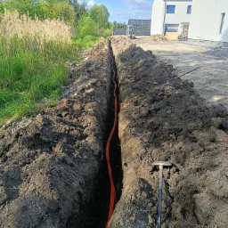 Wykopany rów z pomarańczowymi kablami w ziemi, widok z góry. W tle budynki i zieleń. Obok rowu leży szpadel. Słoneczny dzień.