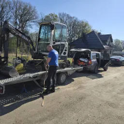 Mini koparka Terex na lawecie, zabezpieczana przez mężczyznę w niebieskiej koszulce. W tle domy i samochody.