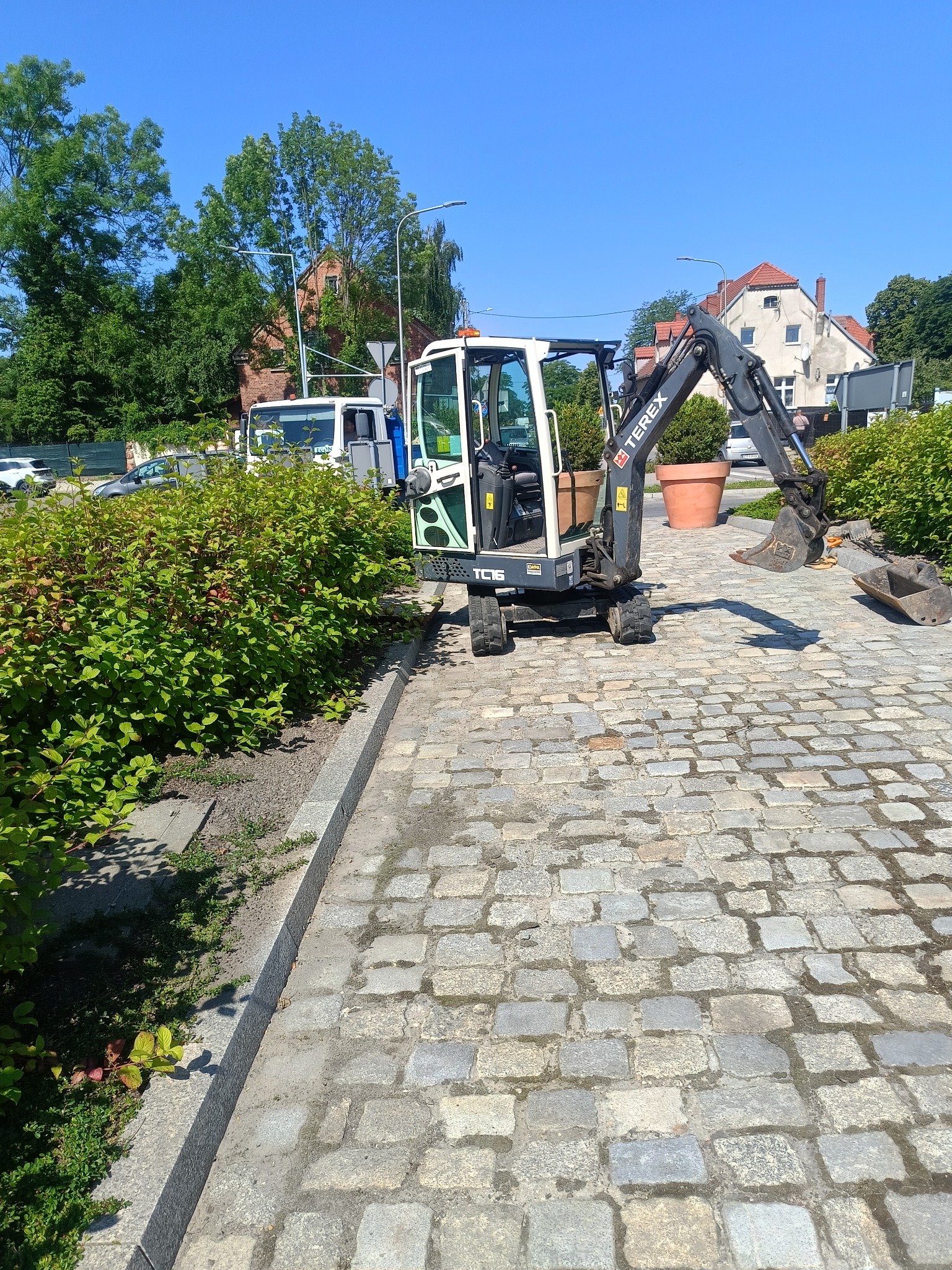 Kompaktowa koparka Terex TC16 na brukowanym placu, z zielenią w tle i budynkami. Widoczny duży doniczki z roślinami. Jasne, słoneczne oświetlenie.