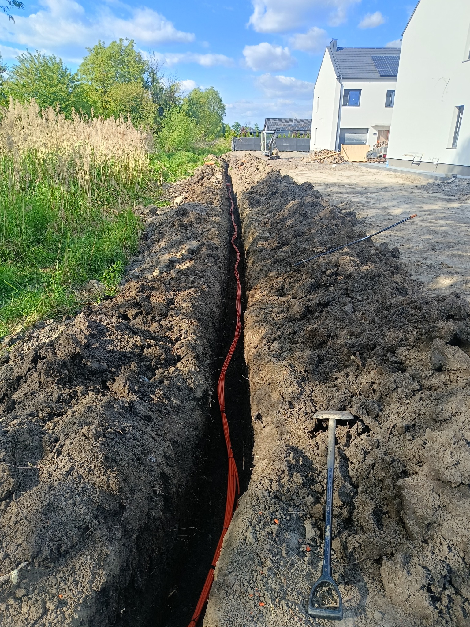 Wykopany rów z pomarańczowymi kablami w ziemi, widok z góry. W tle budynki i zieleń. Obok rowu leży szpadel. Słoneczny dzień.