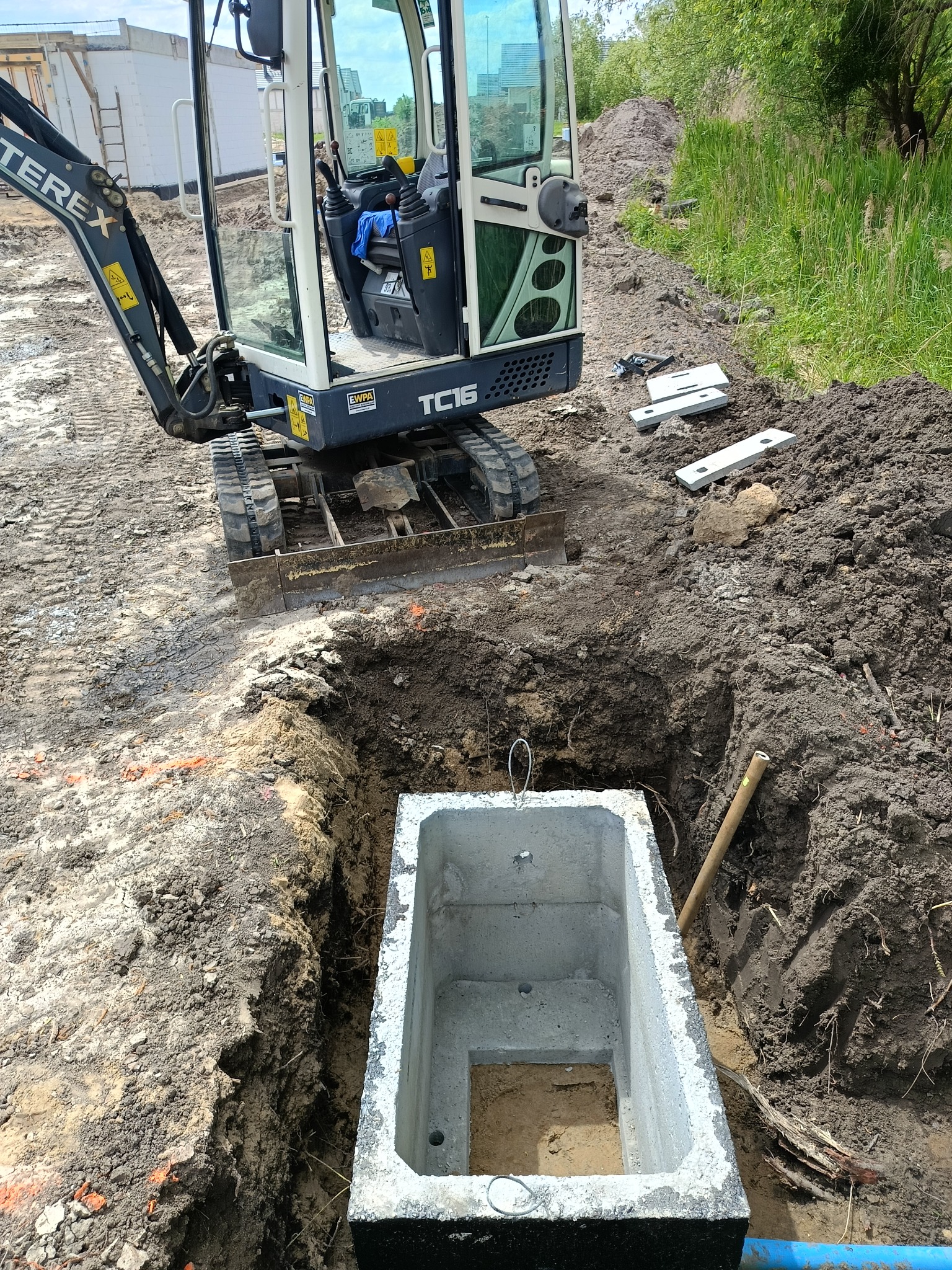 Mini koparka Terex TC16 przy wykopie z betonową studzienką, widoczne płyty betonowe, ziemia i trawa. Prace ziemne w toku, słoneczny dzień.