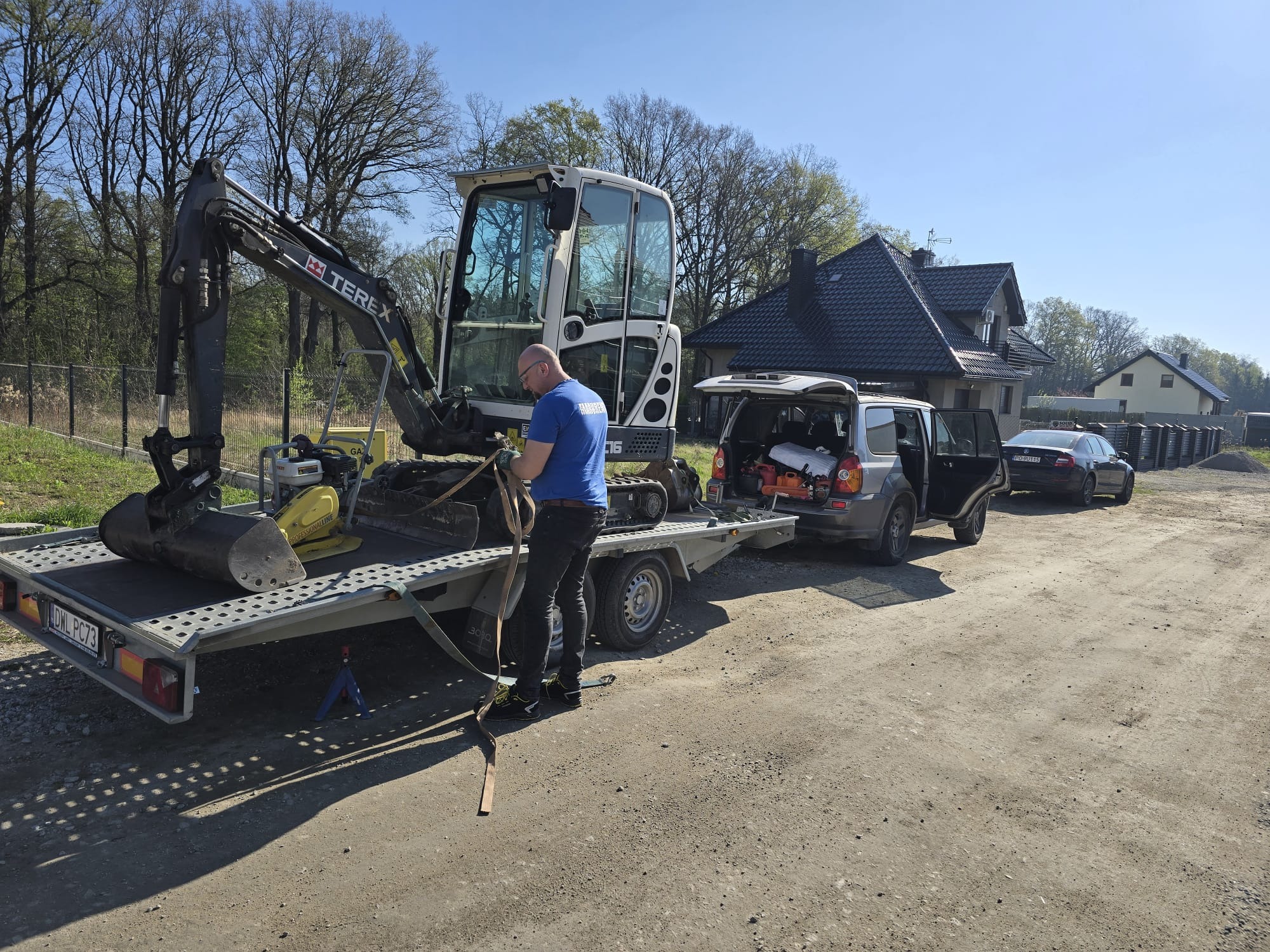 Mini koparka Terex na lawecie, zabezpieczana przez mężczyznę w niebieskiej koszulce. W tle domy i samochody.