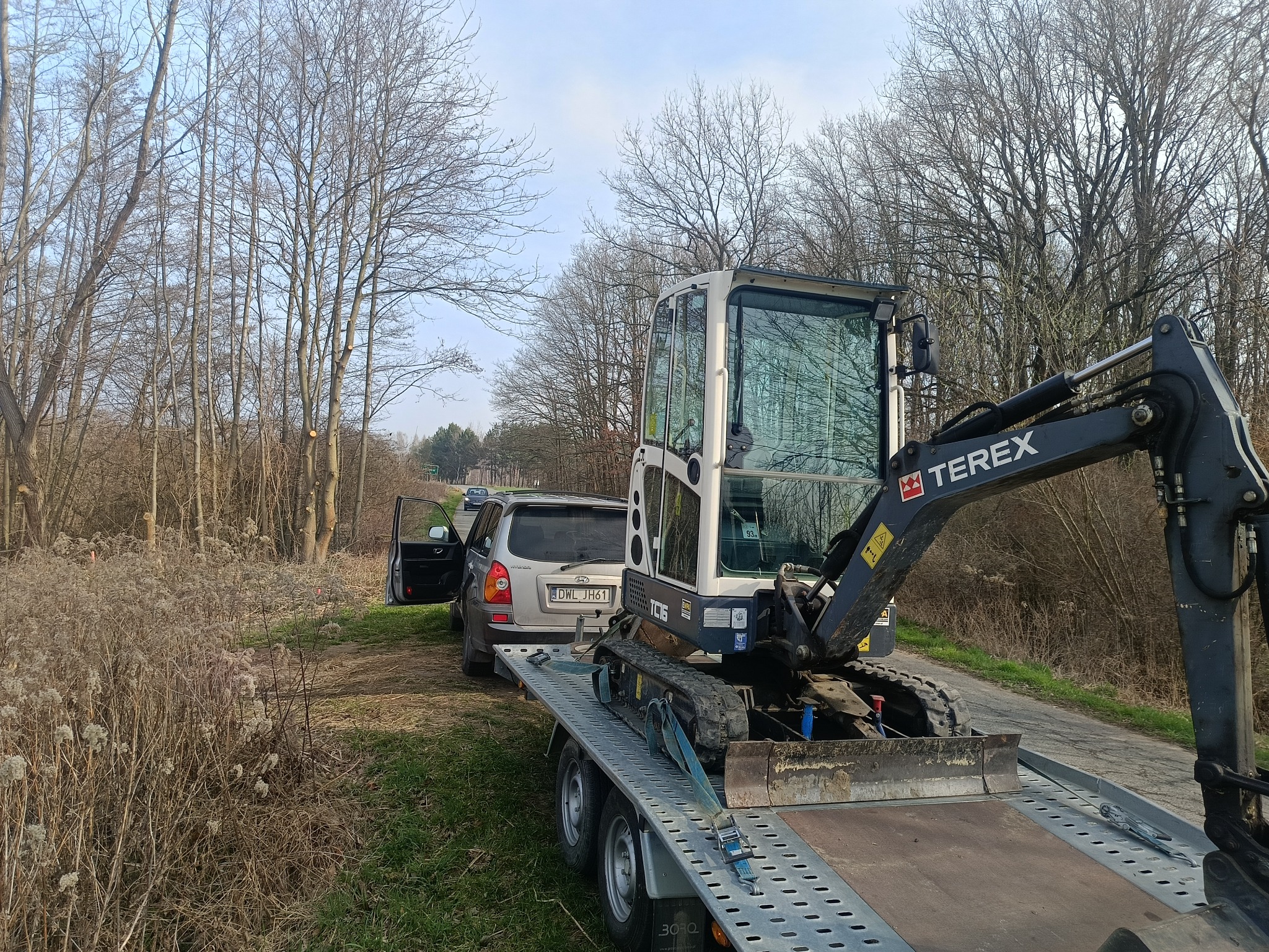 Mini koparka Terex na lawecie, zabezpieczona pasami transportowymi, zaparkowana obok srebrnego auta na poboczu drogi, w tle drzewa i błękitne niebo.