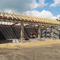 Budowa otwartego garażu z bloczków betonowych z drewnianą konstrukcją dachu, widoczne zbrojenia filarów i podnośnik wewnątrz.