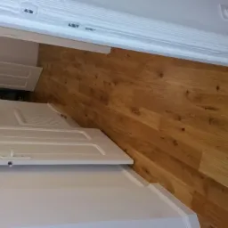 Ułożona podłoga z paneli drewnianych, widoczne białe listwy przypodłogowe i fragmenty białych drzwi z dekoracyjnymi panelami.