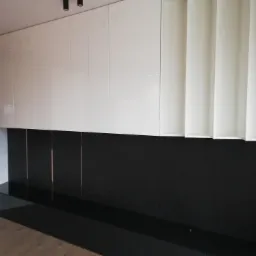 Nowoczesna ściana z szafkami wiszącymi w kolorze białym, połączona z czarnym panelem z podłużnymi wstawkami, oświetlenie punktowe na suficie.