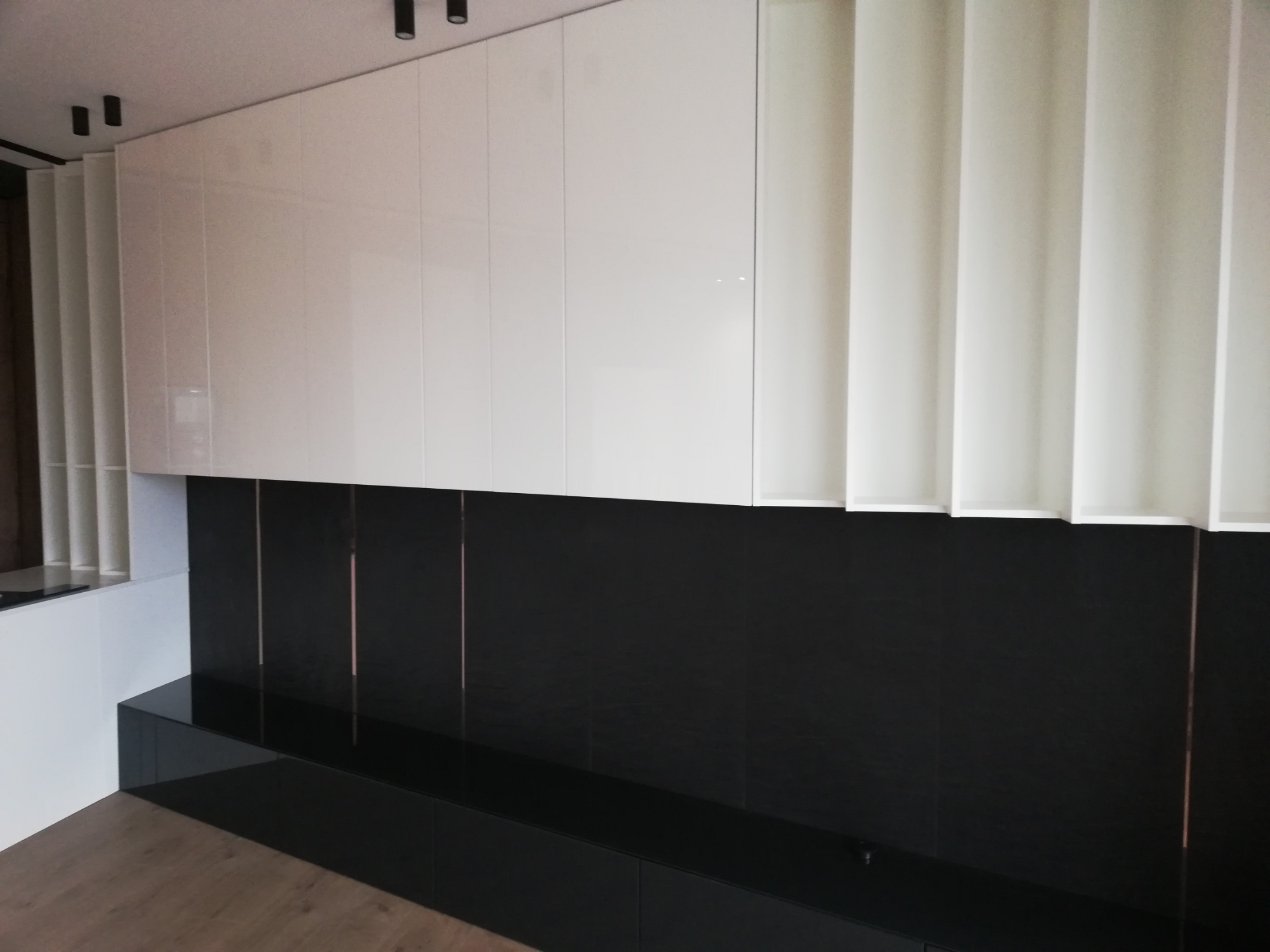 Nowoczesna ściana z szafkami wiszącymi w kolorze białym, połączona z czarnym panelem z podłużnymi wstawkami, oświetlenie punktowe na suficie.