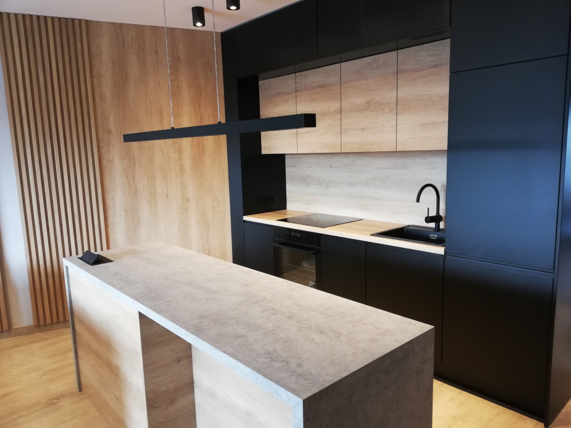 Nowoczesna kuchnia z wyspą: czarne matowe szafki dolne, drewnopodobne szafki górne, betonowy blat wyspy, pionowe lamele na ścianie i podłużna lampa sufitowa.