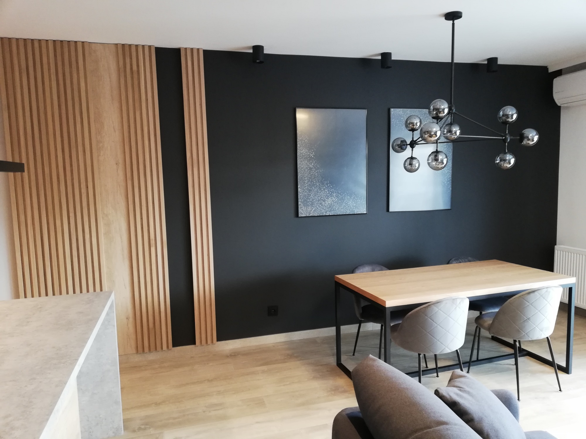 Nowoczesna jadalnia z drewnianym stołem na metalowych nogach, szarymi krzesłami z pikowanym wzorem, czarną ścianą z drewnianymi lamelami i abstrakcyjnymi obrazami oraz designerską lampą sufitową.