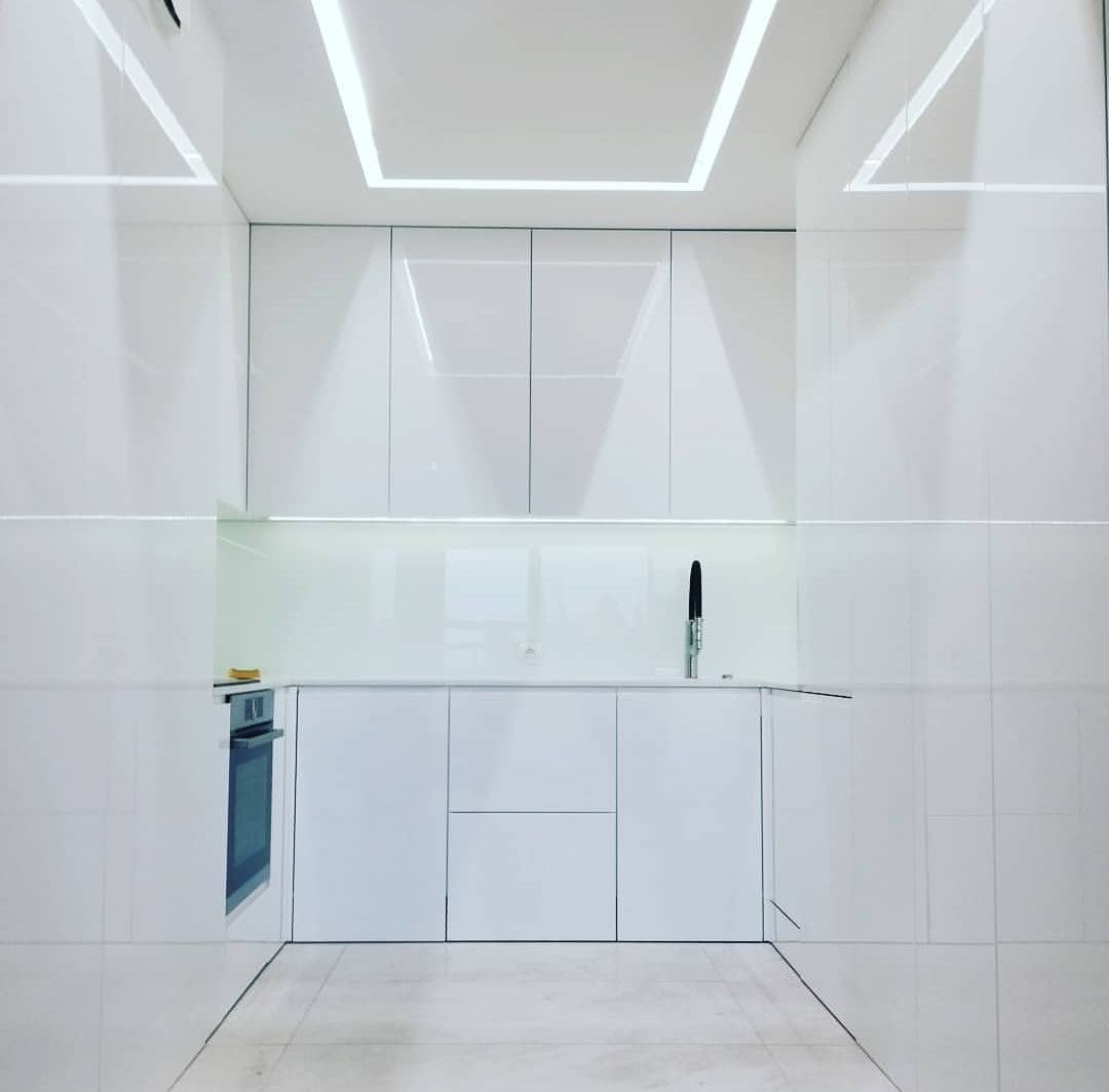 Minimalistyczna, biała kuchnia z błyszczącymi frontami szafek, czarną baterią i oświetleniem LED w suficie, widziana z niskiego kąta.