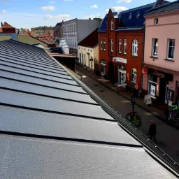 Nowoczesne krycie dachu z blachy na tle miejskiej ulicy z budynkami o zróżnicowanej architekturze i błękitnym niebem z chmurami.
