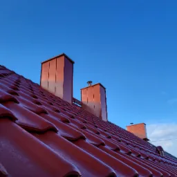 Czerwona dachówka ceramiczna z trzema kominami w kolorze dopasowanym do dachu, widok z dołu na tle błękitnego nieba, widoczna rynna metalowa.