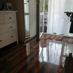 Błyszcząca, ciemna podłoga z paneli, odbijająca światło wpadające przez otwarte drzwi balkonowe z białymi zasłonami, widoczna komoda z szufladami i szafa w tle.