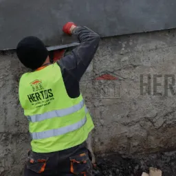 Pracownik w odblaskowej kamizelce firmy HERTOS uszczelnia połączenie betonowej ściany fundamentowej z izolacją przeciwwilgociową.