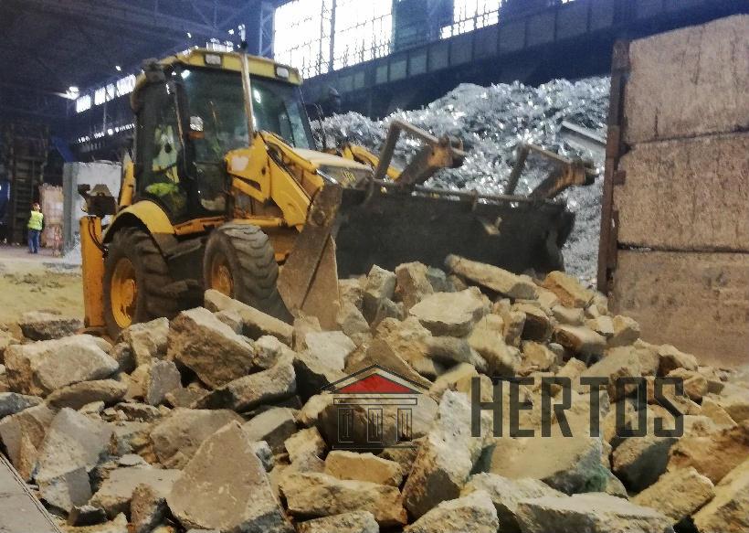 Żółta koparko-ładowarka na tle hali przemysłowej, z przodu sterta gruzu betonowego, w tle pryzma złomu, w oddali widoczna sylwetka pracownika w kamizelce odblaskowej.