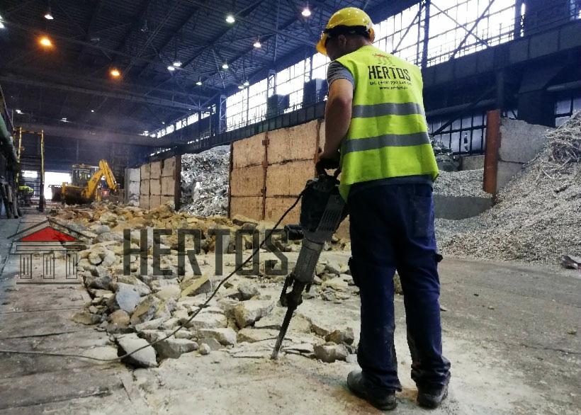 Pracownik w żółtym kasku i kamizelce używa młota pneumatycznego do rozbijania betonowej posadzki wewnątrz hali, w tle widoczny stos gruzu i koparka.