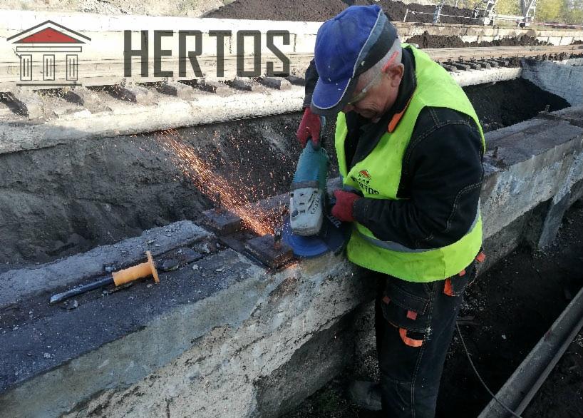 Pracownik w kamizelce odblaskowej szlifuje metalowy element w prefabrykacie betonowym, iskry rozbryzgują się wokół, obok leży przecinak z żółtą rączką.