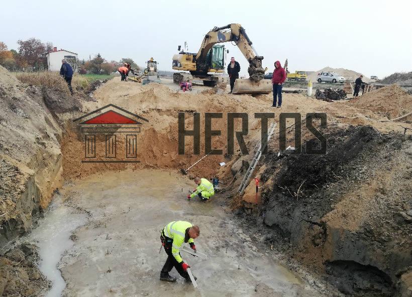 Przygotowanie wykopu pod fundamenty: dwóch pracowników w jaskrawych kamizelkach mierzy głębokość, w tle koparka i obserwujący robotników mężczyźni.