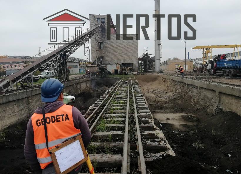 Geodeta w pomarańczowej kamizelce z napisem 'GEODETA' i niebieskim kasku, trzymający clipboard z planami, stoi obok torów kolejowych na tle budowy hali z podajnikiem taśmowym i pracującym dźwigiem.