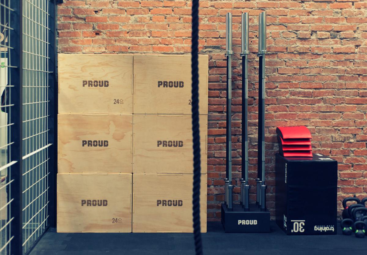 Wnętrze siłowni z ceglaną ścianą, drewnianymi skrzyniami z napisem 'PROUD', stojakami na sztangi i czarnymi skrzyniami do ćwiczeń.