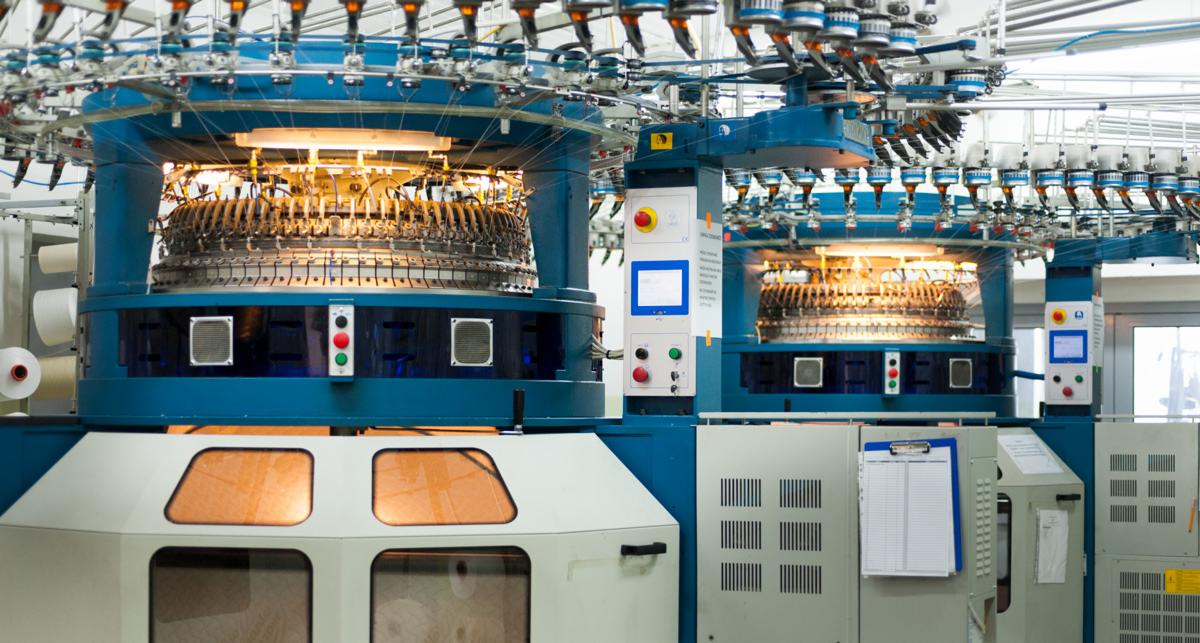 Wielkoformatowa, automatyczna maszyna dziewiarska z centralnym cylindrem igłowym, widoczne liczne prowadnice nici i panele sterowania z przyciskami oraz ekranami.