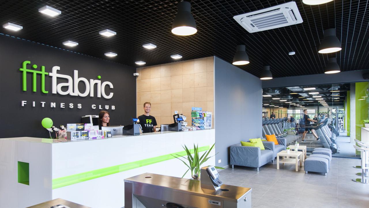 Wnętrze nowoczesnego klubu fitness Fit Fabric z recepcją, logo na ścianie, strefą relaksu i widocznymi bieżniami w tle.