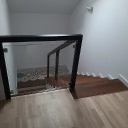 Klatka schodowa z drewnianymi schodami i szklaną balustradą z czarnymi elementami, widok z g&oacute;ry na d&oacute;ł, z widoczną podłogą z paneli i wzorzystymi płytkami na podeście.