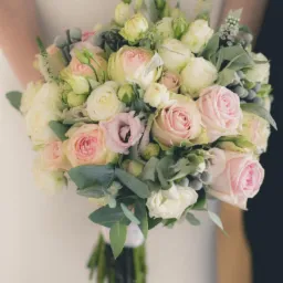 Delikatny bukiet ślubny z różowych i białych róż oraz eukaliptusa, trzymany przez osobę w białej sukience, na ciemnym tle.