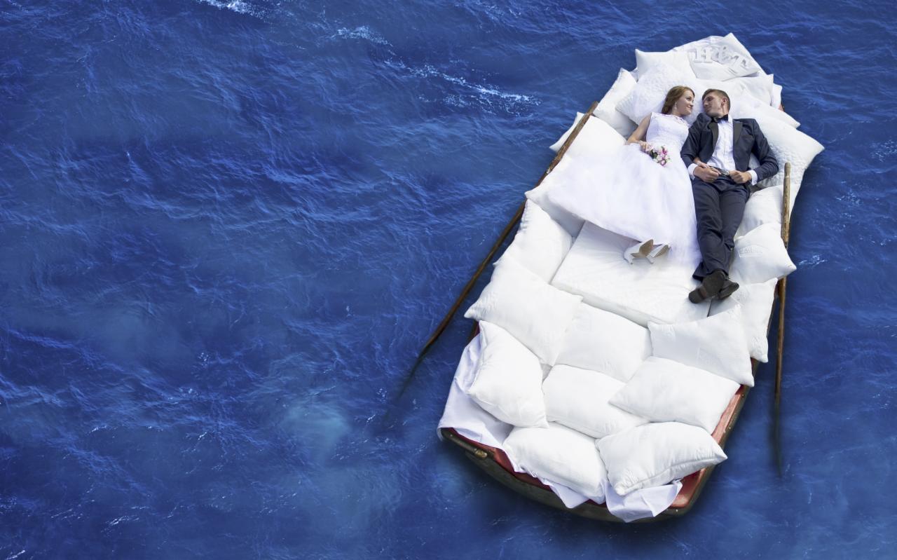 Nowożeńcy leżący na łodzi wypełnionej białymi poduszkami, dryfującej po ciemnoniebieskim oceanie, panna młoda w białej sukni i bukiecie, pan młody w ciemnym garniturze, ujęcie z góry.