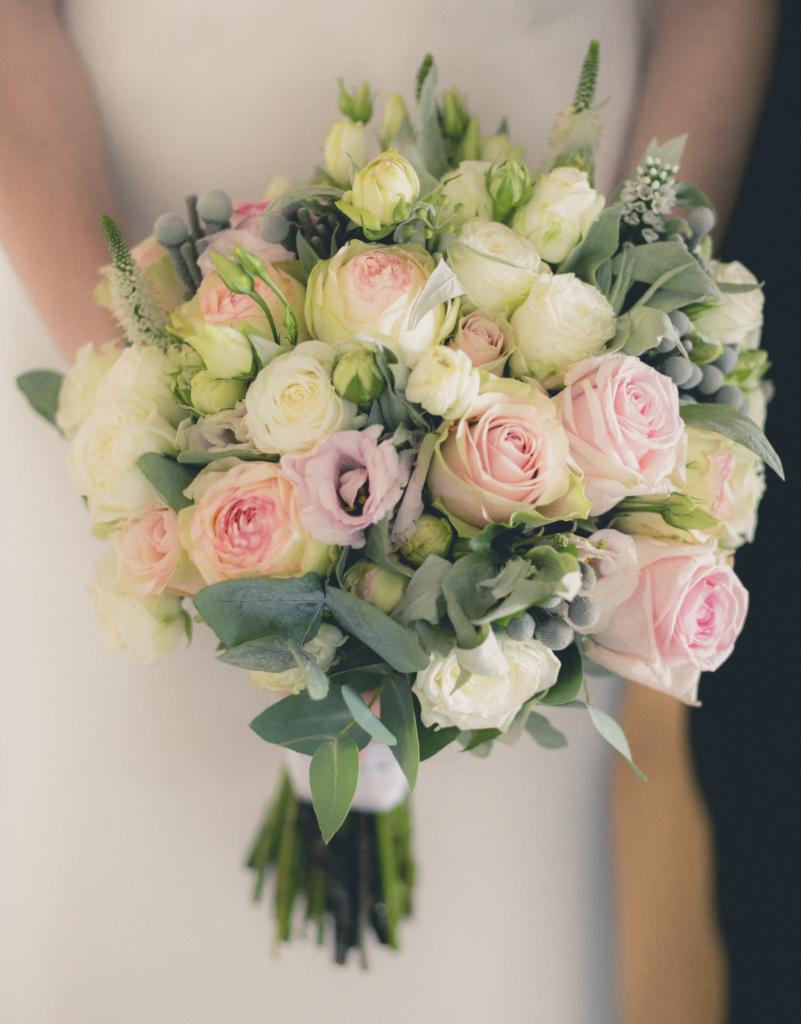 Delikatny bukiet ślubny z różowych i białych róż oraz eukaliptusa, trzymany przez osobę w białej sukience, na ciemnym tle.