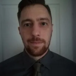 Portret mężczyzny z rudą brodą w szarej koszuli i krawacie, z delikatnym uśmiechem, na tle białych drzwi.