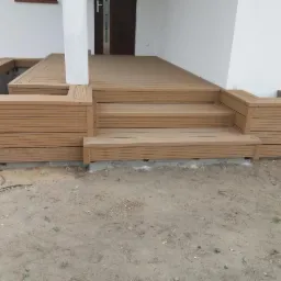 Jasny taras z desek kompozytowych z trzema schodami prowadzącymi do drzwi wejściowych, otoczony niskimi ściankami z tego samego materiału, widok z poziomu gruntu na etapie prac wykończeniowych.