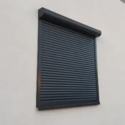 Zamknięta, czarna roleta zewnętrzna na jasnej, teksturowanej ścianie budynku, widok z dołu.