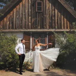 Para młoda w plenerze przed drewnianym domem. Suknia z rozkloszowanym dołem, delikatne zdobienia. Pan młody w białej koszuli i czarnych spodniach.